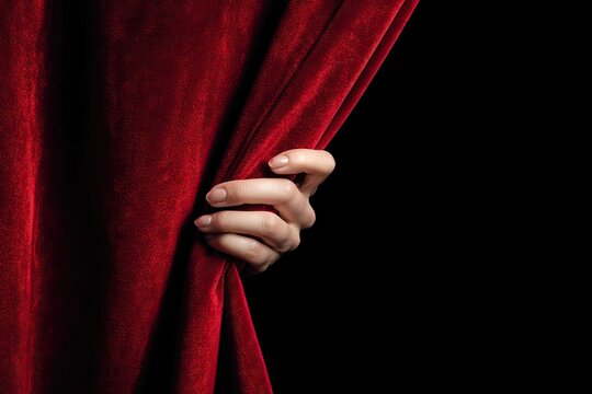 A hand pulls back a luxurious red velvet stage curtain, revealing a dark void