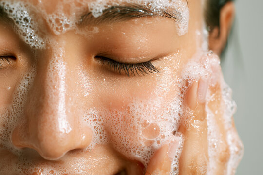 Close-up of a young woman washing her face with a foaming cleanser. Concept of daily skincare routine, facial hygiene, and beauty treatment featuring soft bubbles and clean skin