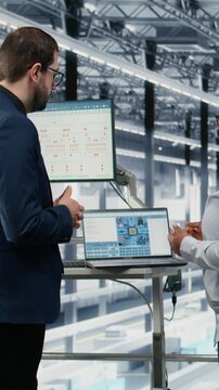 Vertical video Female manager and coworker review microchip factory diagnostic readouts to prevent downtime. Management executives study semiconductor manufacturing plant data, doing quality control