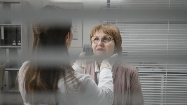 Medium shot through medical office blinders of female physician palpating lymph nodes of elderly woman then measuring temperature with digital thermometer during routine physical examination