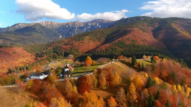 Aerial view of colorful autumn trees and a small village nestled in the Carpathian Mountains with high peaks in the background in Bran, Brasov, Romania.