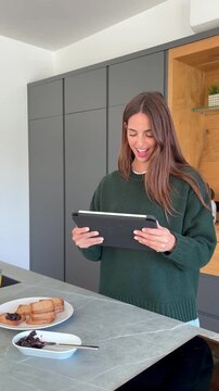 A cheerful young woman in a cozy sweater engages with her digital tablet in a modern kitchen. She smiles while browsing, with breakfast items like rusks and jam ready on the counter, depicting a relax