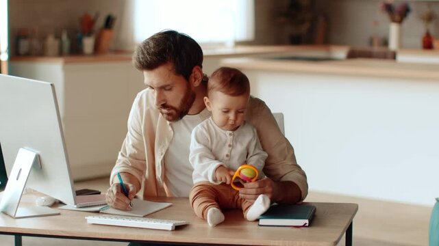 A man with light skin is focused on his work at a desk in a bright kitchen. A baby sits on his lap, playing with a toy while he writes notes. The atmosphere is warm and inviting.