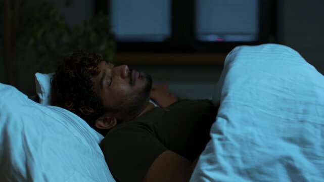 A young man with curly hair rests comfortably in bed, covered with white sheets. The room has soft lighting that highlights his peaceful expression during the night.