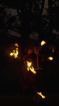 Mystical fire performer in ornate costume spinning blazing torches at night in a dark forest, creating trails of light and dramatic sparks in the shadows