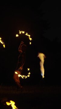 Nighttime fire dancer performing dynamic spinning choreography with blazing torches, creating dramatic light trails and glowing patterns against a dark outdoor backdrop