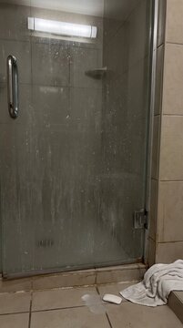 Dirty glass shower door covered in soap scum buildup with a dropped bar soap and towel on the tile floor