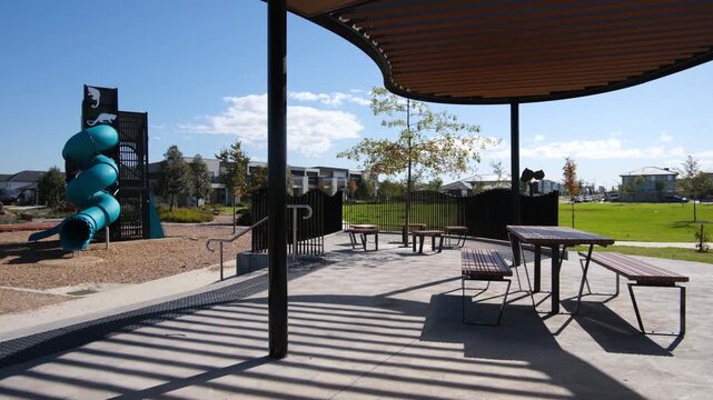 A modern community park in a new outer suburban housing estate in Tarneit, Melbourne, Australia, featuring a playground with wood chip softfall, shade shelter with picnic tables and bench seating
