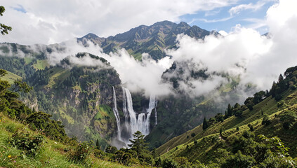 Obraz premium Majestic waterfall cascades down steep mountainside. Clouds drift between rugged, green-hued peaks. Lush vegetation blankets rolling hillsides. Distant cliffs frame the powerful, misty cascade