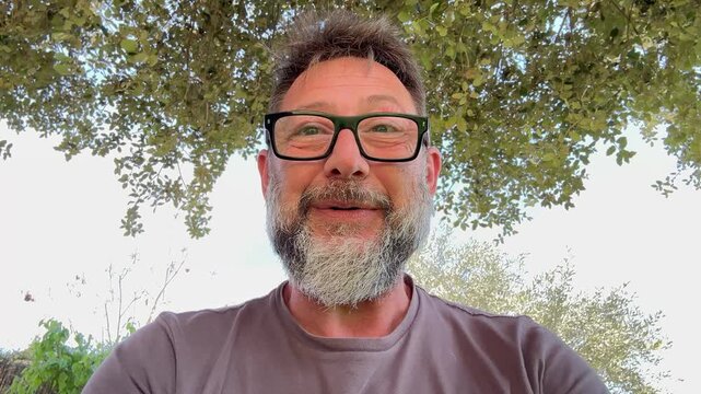 A happy bearded man wearing glasses smiles genuinely while standing outdoors under green tree leaves. He appears relaxed and cheerful in natural daylight.