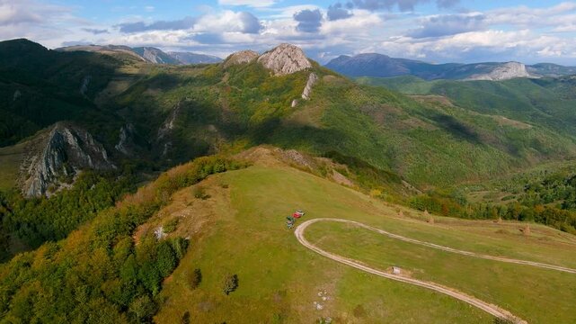 Aerial view of a winding dirt road on a grassy ridge overlooking rocky mountain peaks and lush forests in Salciua de Jos, Alba County, Romania.
