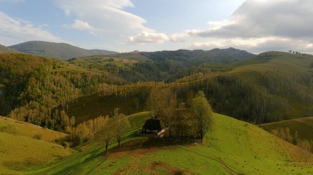 Aerial view of a traditional wooden house on a green hill ridge surrounded by autumn forest and mountains in Salciua de Jos, Alba County, Romania.