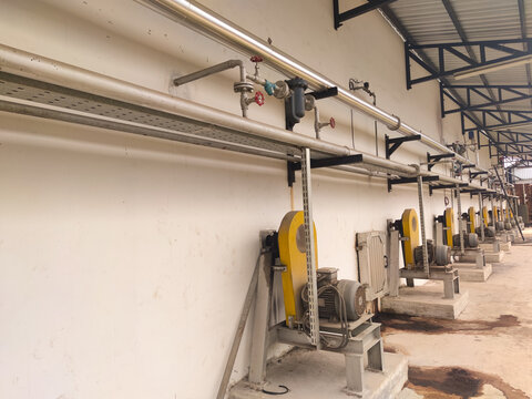 Row of industrial exhaust blowers with yellow guards and overhead utility pipes on a factory wall. Professional engineering infrastructure for manufacturing plant ventilation