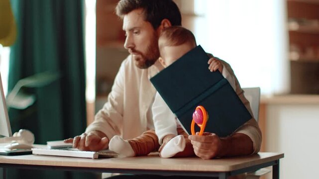 A man with light skin is seated at a desk working on a computer while a baby is playing with a colorful toy next to him. The setting is bright and full of natural light.