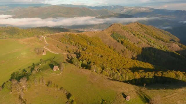 Aerial view of traditional wooden huts scattered across green hills and autumn forests with low clouds in the valley of Salciua de Jos, Alba County, Romania.