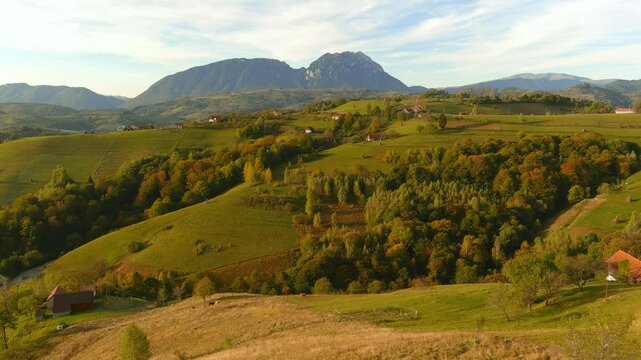 Aerial view of rolling green hills with autumn foliage and scattered houses against a mountain backdrop in Bran, Brasov, Romania.