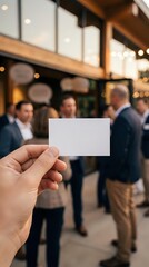 Obraz premium Hand holding blank white business card at corporate networking event with blurred professionals in background. Mockup template for branding and contact information.