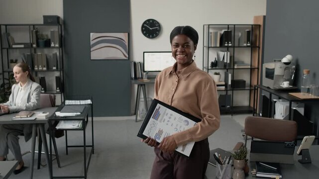 Portrait shot of young African American woman smiling for camera while posing with business documents in modern office