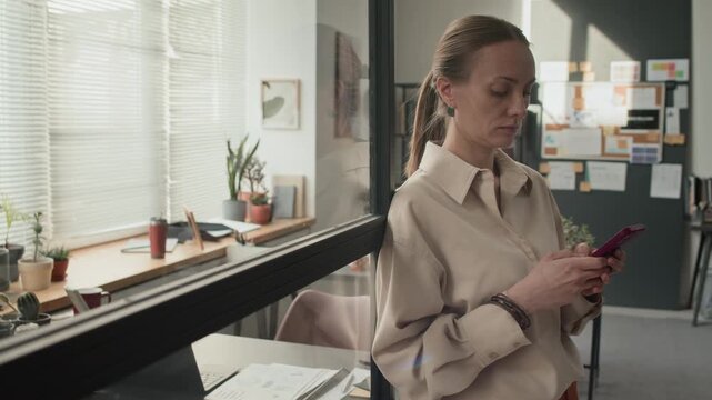 Middle aged Caucasian woman leaning on glass partition wall and texting on her smartphone in modern office