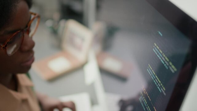 High angle close up shot of young African American woman in glasses looking at screen while writing code in office