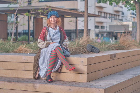 Redheaded woman in colorful bohemian outfit making silence gesture on city bench