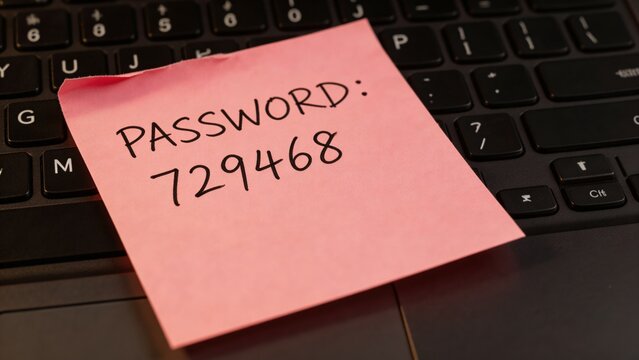A close-up of a pink sticky note with a handwritten password on a black laptop keyboard, highlighting the risks of insecure password storage and potential cyber threats.