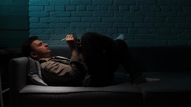 young man lying on a dark couch, holding a pen and notebook, experiencing creative thought and inspiration under a blue light shining on a brick wall