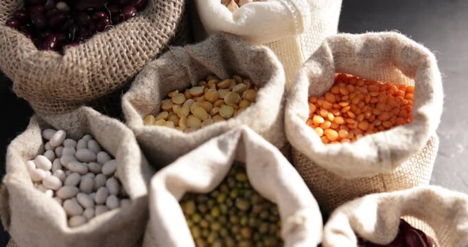 Different types of legumes in burlap bags on black table, above view. Camera moving right and down