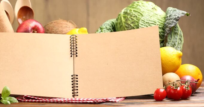Open recipe book, kitchen utensils and fresh products on wooden table, closeup. Camera moving right