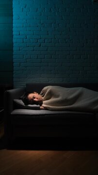 Vertical video man laying down on a sofa at night, unable to sleep, experiencing insomnia and suffering from a restless mind in a dark room with brick wall