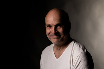 Obraz premium Mature bald man with a beard smiling against a dark studio background, wearing a white t shirt