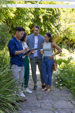Diverse coworkers standing on stone path under trellis discussing plants, making notes on clipboard