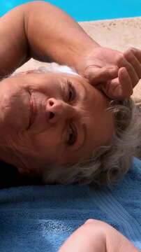 Vertical video: Shielding senior woman lying on blue pool towel, reacting to sunlight in swim top
