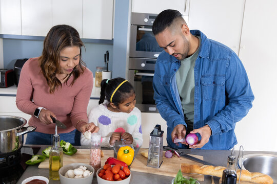 Family of three chopping vegetables and seasoning food at kitchen island with cutting boards