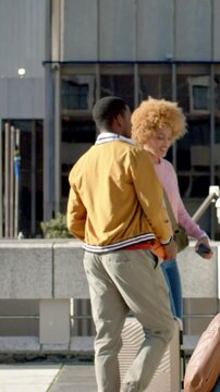 Vertical video: Walking in sun plaza, man in jacket with basketball and case, woman checking phone