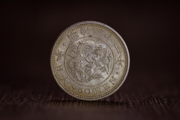 Close-up photo of a Japanese 50 sen silver coin from the Meiji era, shown as a single front view standing upright on a dark textured surface. © Mr