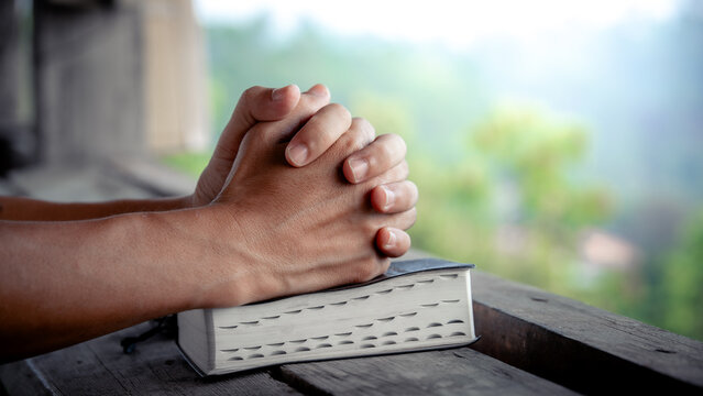 Close up of a man praying, hands clasped together on Bible, devotional concept.