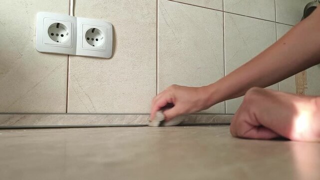 Low angle - a woman wipes the baseboard and floor by the kitchen wall next to the outlet, home cleaning, cleanliness, hygiene, interior care.
