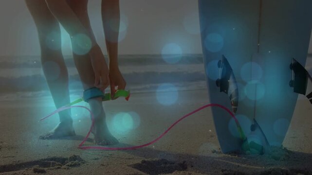 Woman shifting weight by board, grabbing cuff, fastening leash with blue bokeh, preparing to surf