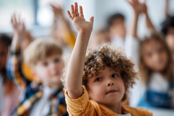 Fototapeta premium Curly-haired child raises hand eagerly. Bright eyes show curiosity and excitement. Classroom setting with blurred peers behind. Ideal for education, learning, or child engagement themes