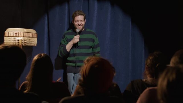 Young male stand up comic standing on stage at comedy club, holding microphone and speaking to audience during humorous performance