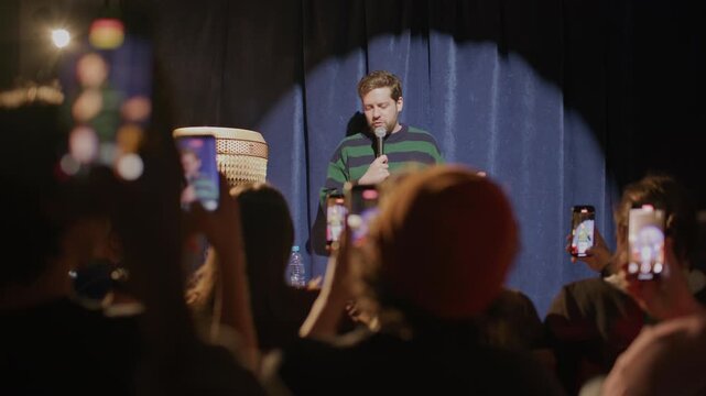 Male comedian performing stand up routine under spotlight on stage as audience capturing his live show with mobile phones