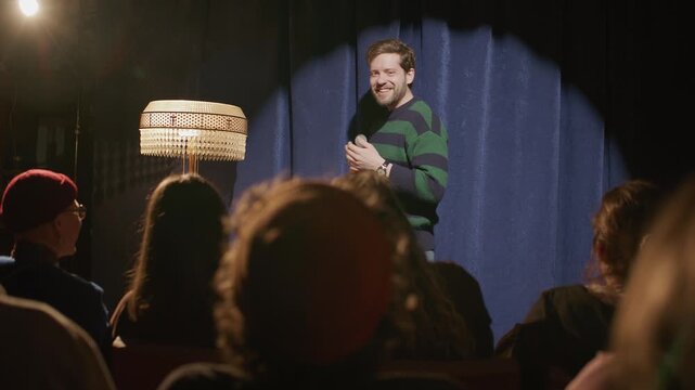 Male comedian holding microphone while performing stand up comedy under spotlight on stage in front of seated audience, delivering humorous monologue during live set