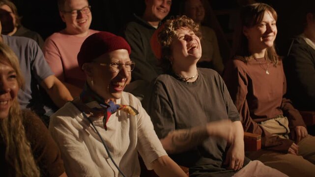 Group of joyful spectators laughing at jokes of comedian during his live stand up performance on stage at comedy club