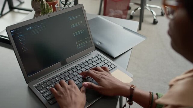 High angle over the shoulder close up shot of African American woman in glasses writing code on laptop, sitting at table in modern office