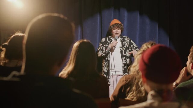 Young female standup comedian in stylish outfit and orange beanie walking on stage, greeting audience and starting her monologue under spotlight during open mic night