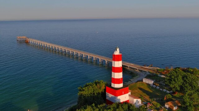 Aerial view of the red and white striped Shabla Lighthouse and a long pier extending into the Black Sea at sunset in Bulgaria.