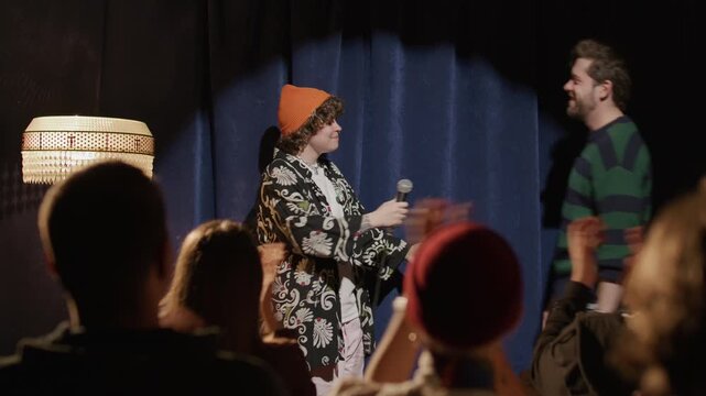 Young female host welcoming guest comedian on stage at stand up club, he taking microphone and excitedly greeting audience as beginning his performance