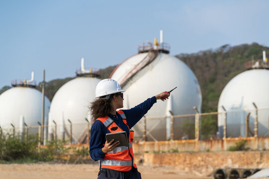 Female environmental engineer in safety vest and helmet using radio and tablet at industrial gas storage tanks for energy and fuel supply operations.