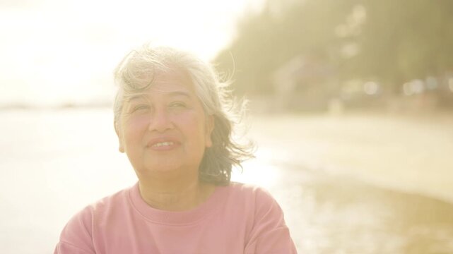 Happy Asian senior woman smiling at beach during sunset, enjoying peaceful moment, active elderly lifestyle, carefree summer holiday vacation, aging gracefully healthy retirement relaxing by seaside.
